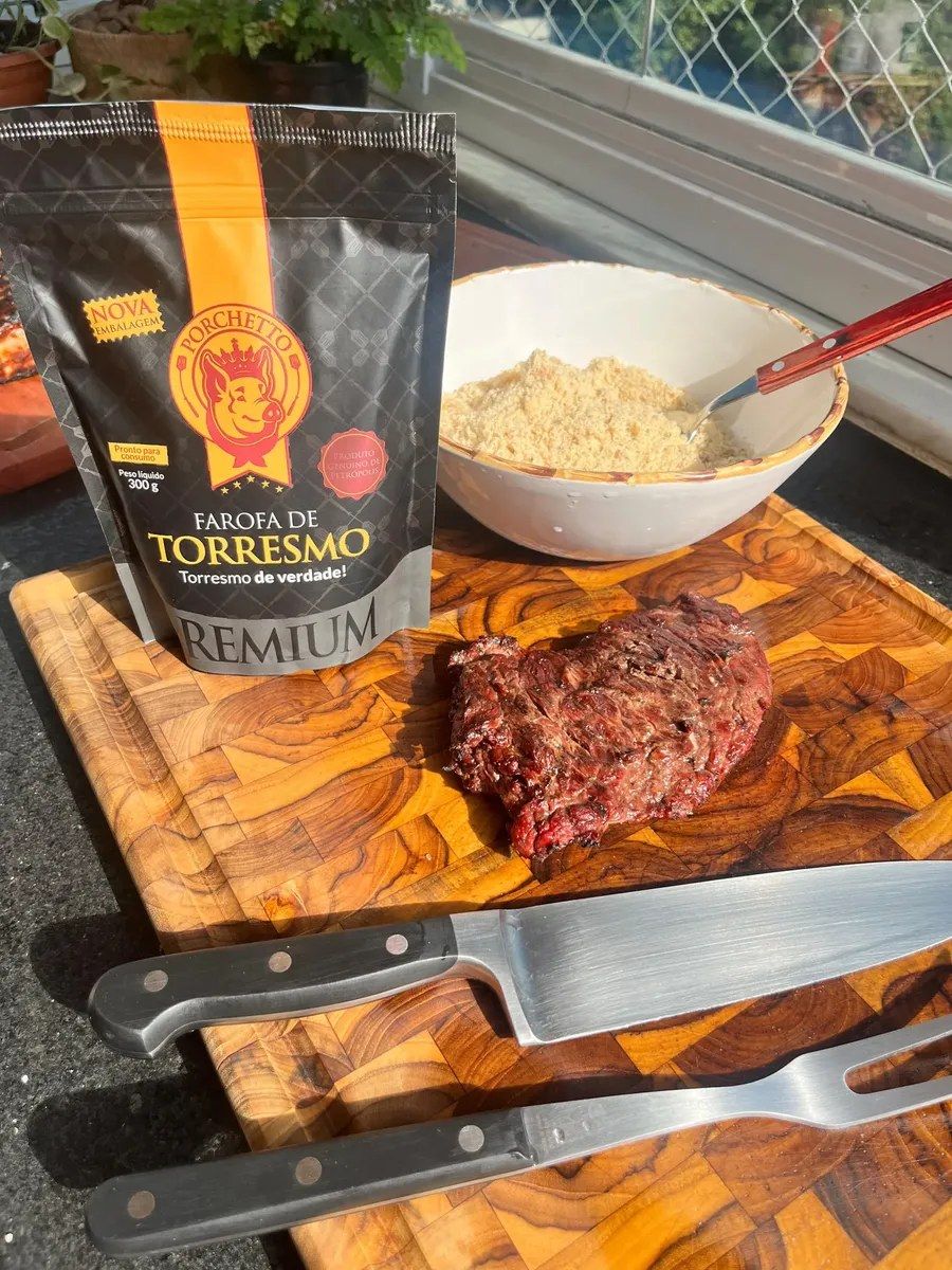 Pacote da farofa Porchetto ao lado de uma tigela de farofa, carne grelhada na tábua e talheres de churrasco